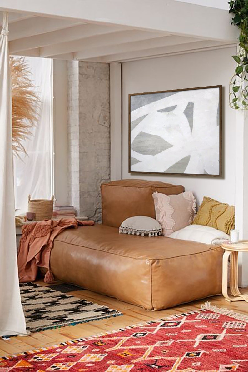 May include: A brown leather couch with white and beige pillows in a living room with a white and gray abstract painting on the wall. The couch is in front of a window with a sheer curtain and a basket on the windowsill. There is a red and orange rug on the floor.
