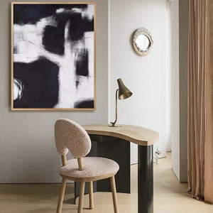May include: A black and white abstract painting hangs above a wooden desk with a curved top. A fluffy white chair sits in front of the desk. A gold lamp with a white shade sits on the desk.