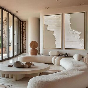Op de afbeelding: Twee abstracte beige en witte textuur canvas schilderijen met gouden lijsten die aan een muur hangen in een moderne woonkamer met een witte gebogen bank en een ronde salontafel.
