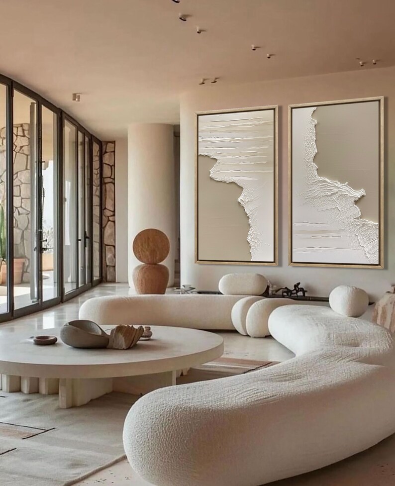 Op de afbeelding: Twee abstracte beige en witte textuur canvas schilderijen met gouden lijsten die aan een muur hangen in een moderne woonkamer met een witte gebogen bank en een ronde salontafel.