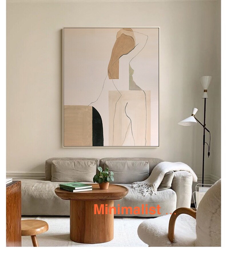 May include: A minimalist living room featuring a beige sofa, a wooden coffee table, and a framed abstract painting depicting a nude figure in shades of beige and brown. The painting is positioned above the sofa, with the coffee table situated in front. The coffee table is round and has a wooden base. The painting is framed in a thin, light wood frame. The sofa is upholstered in a light beige fabric. The room is decorated in a minimalist style with a neutral colour palette.