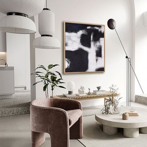 May include: A modern living room interior with a black and white abstract painting hanging on a white wall. A brown velvet armchair sits in front of a concrete coffee table with a vase of dried flowers.