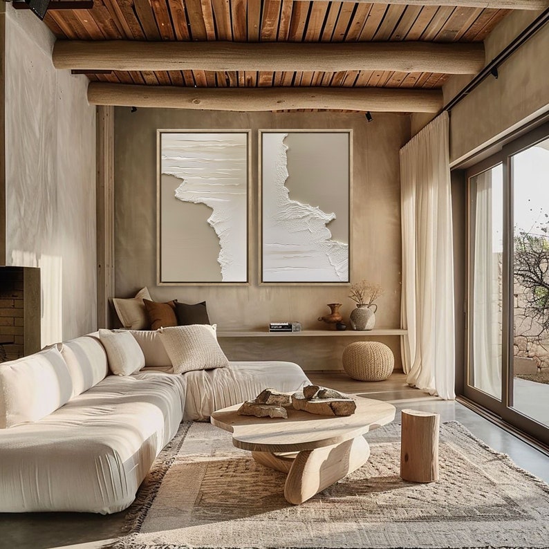 Op de afbeelding: Een moderne woonkamer met een witte modulaire bank, een houten salontafel en twee grote abstracte schilderijen in beige en witte tinten. De kamer heeft een houten plafond en grote ramen met witte gordijnen.