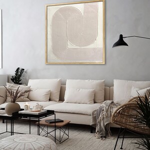 Peut inclure: Un canap&eacute; blanc avec une couverture et un pouf ottoman blanc devant un mur gris clair avec une peinture abstraite encadr&eacute;e en rose et blanc. La peinture pr&eacute;sente une s&eacute;rie de lignes courbes et est encadr&eacute;e dans un cadre en bois naturel.