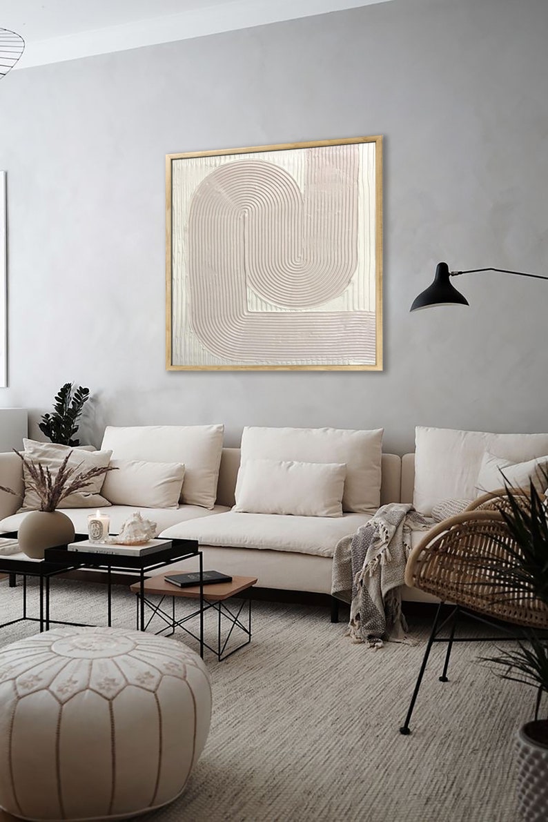 Peut inclure: Un canap&eacute; blanc avec une couverture et un pouf ottoman blanc devant un mur gris clair avec une peinture abstraite encadr&eacute;e en rose et blanc. La peinture pr&eacute;sente une s&eacute;rie de lignes courbes et est encadr&eacute;e dans un cadre en bois naturel.