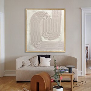 Peut inclure: Une peinture abstraite rose p&acirc;le avec un design textur&eacute; et superpos&eacute; dans un cadre dor&eacute;. La peinture est accroch&eacute;e &agrave; un mur dans un salon avec un canap&eacute; beige, un pouf brun, une plante en pot et une table d'appoint avec une lampe.
