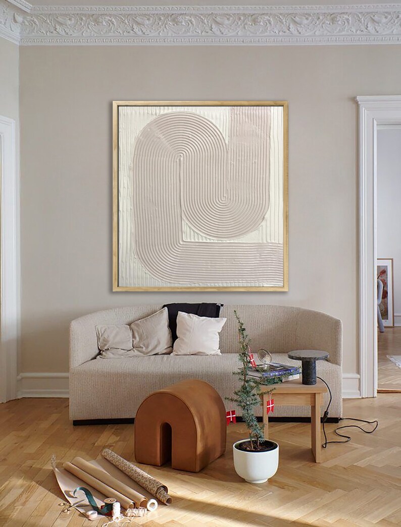 Peut inclure: Une peinture abstraite rose p&acirc;le avec un design textur&eacute; et superpos&eacute; dans un cadre dor&eacute;. La peinture est accroch&eacute;e &agrave; un mur dans un salon avec un canap&eacute; beige, un pouf brun, une plante en pot et une table d'appoint avec une lampe.