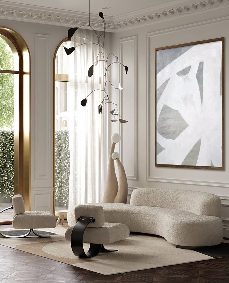 May include: A living room interior with a white, curved sofa, two white chairs with black metal bases, a white and gray abstract painting, and a black mobile hanging from the ceiling.