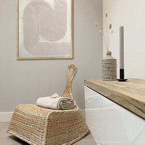 Peut inclure: Une chaise ber&ccedil;ante en osier tress&eacute; avec une finition brun clair. La chaise est pos&eacute;e sur un sol en carrelage brun clair. Une serviette blanche est pli&eacute;e sur l'assise de la chaise. Un meuble blanc avec un dessus en bois se trouve en arri&egrave;re-plan. Un vase avec des fleurs s&eacute;ch&eacute;es et une bougie sont sur le meuble.
