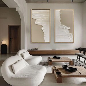 Op de afbeelding: Twee abstracte beige en witte ingelijste canvas schilderijen met een textuur, gelaagde look, hangend aan een muur boven een houten plank en twee witte, gebogen banken in een moderne woonkamer.