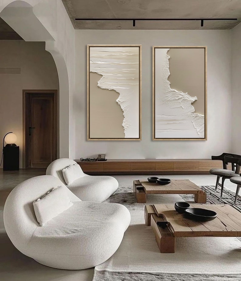 Op de afbeelding: Twee abstracte beige en witte ingelijste canvas schilderijen met een textuur, gelaagde look, hangend aan een muur boven een houten plank en twee witte, gebogen banken in een moderne woonkamer.