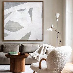 May include: A white and gray abstract painting hangs above a beige sofa and a white sheepskin chair in a living room. The painting is in a wooden frame. The sofa has a white throw blanket on it. The chair has wooden legs. There is a wooden coffee table in front of the sofa.