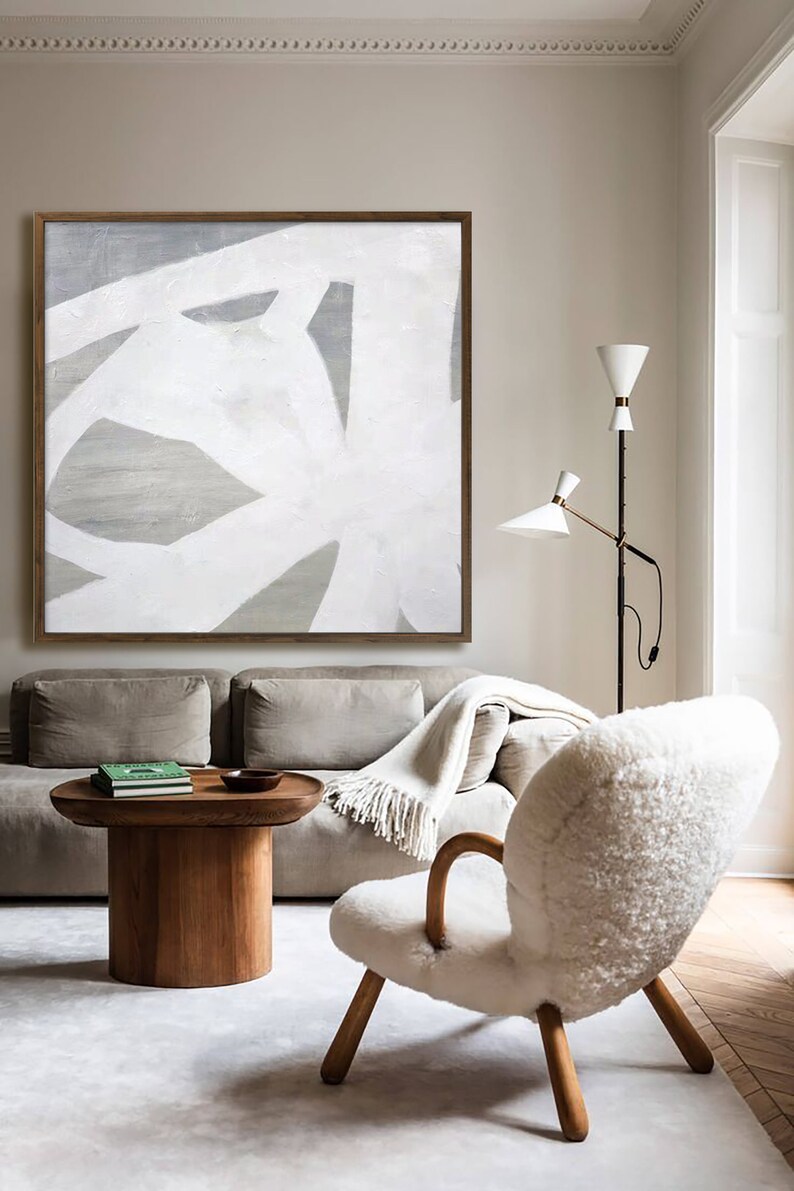 May include: A white and gray abstract painting hangs above a beige sofa and a white sheepskin chair in a living room. The painting is in a wooden frame. The sofa has a white throw blanket on it. The chair has wooden legs. There is a wooden coffee table in front of the sofa.