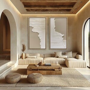 Op de afbeelding: Twee abstracte beige en witte ingelijste canvas wandkunstwerken met een textuurrijk, gelaagd ontwerp. De kunst hangt aan een muur in een woonkamer met een beige hoekbank en een houten salontafel.