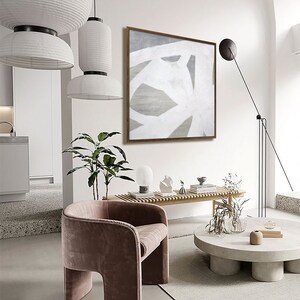 May include: A modern living room interior with a white and gray abstract painting hanging on the wall above a wooden bench. A pink velvet armchair sits in front of a round concrete coffee table with a vase of dried flowers and a book on top.