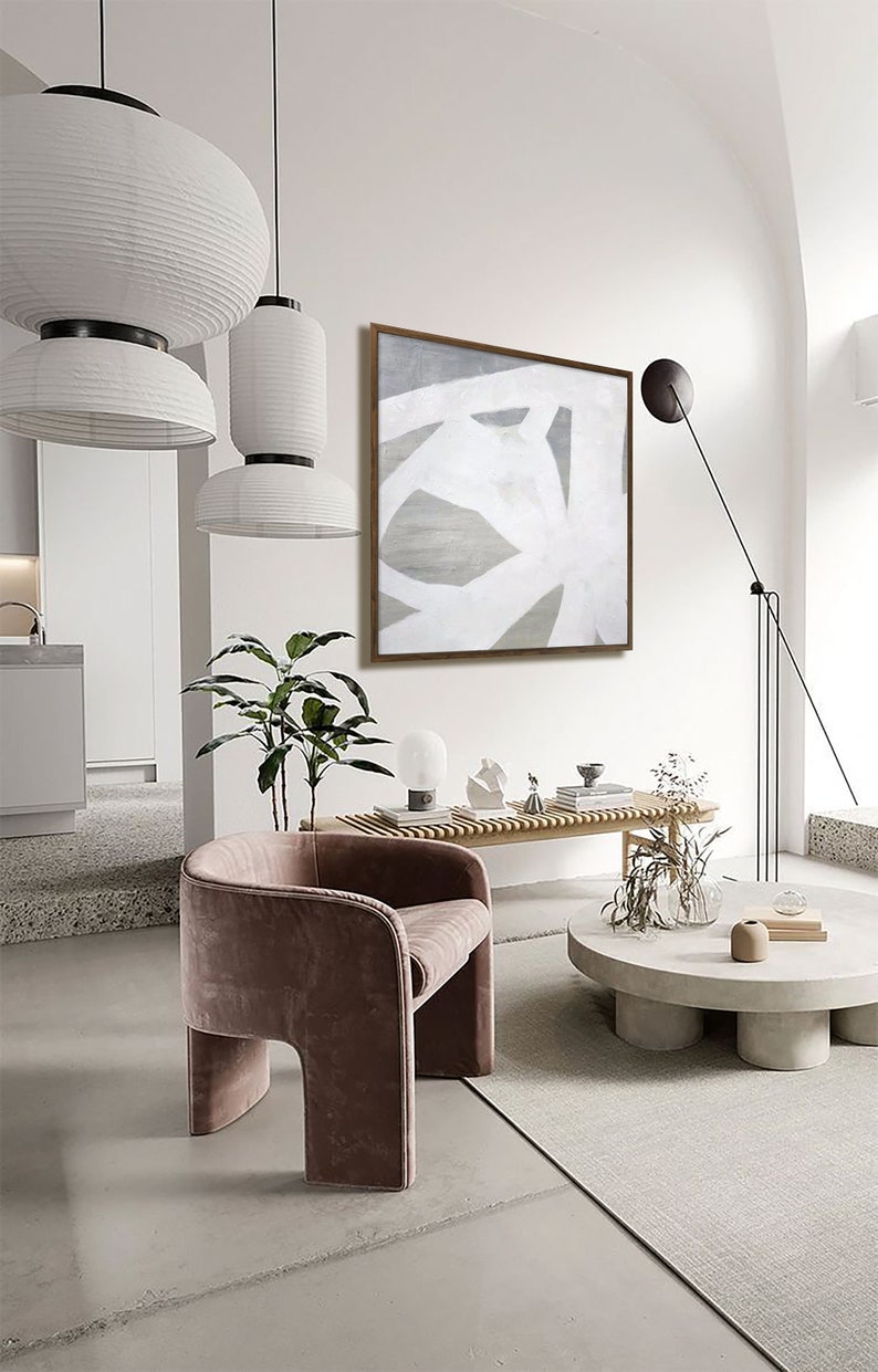 May include: A modern living room interior with a white and gray abstract painting hanging on the wall above a wooden bench. A pink velvet armchair sits in front of a round concrete coffee table with a vase of dried flowers and a book on top.