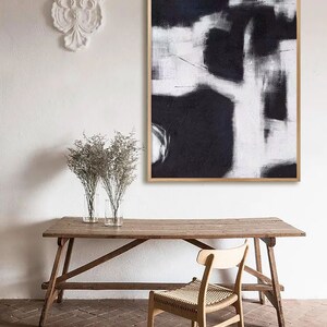 May include: A black and white abstract painting in a wooden frame hangs on a white wall above a wooden table and chair. The painting features bold strokes and a minimalist design.
