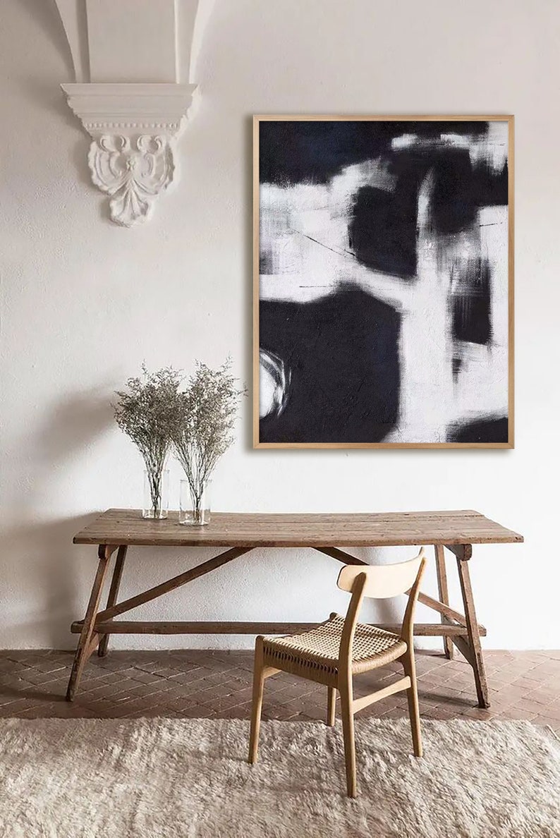 May include: A black and white abstract painting in a wooden frame hangs on a white wall above a wooden table and chair. The painting features bold strokes and a minimalist design.