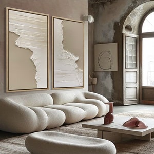 Op de afbeelding: Een witte, pluche, modulaire bank met een lage, witte, ovale poef ervoor. De bank en de poef staan in een woonkamer met een beige kleed, een witte salontafel en twee abstracte schilderijen aan de muur.