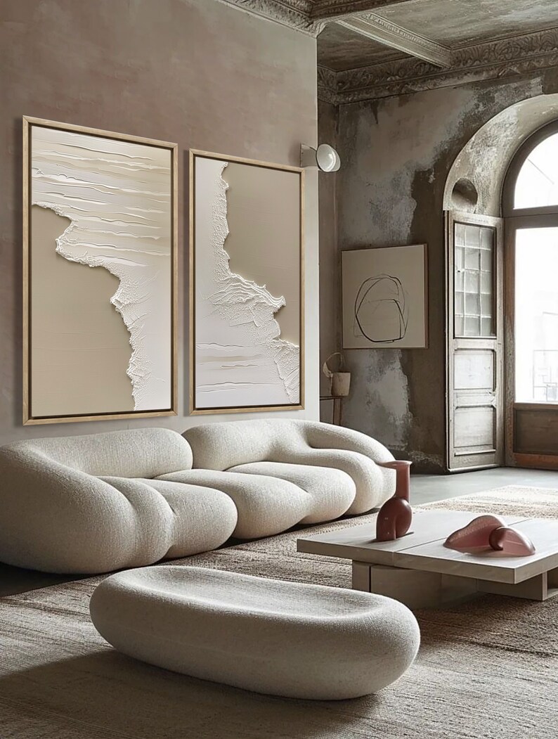 Op de afbeelding: Een witte, pluche, modulaire bank met een lage, witte, ovale poef ervoor. De bank en de poef staan in een woonkamer met een beige kleed, een witte salontafel en twee abstracte schilderijen aan de muur.