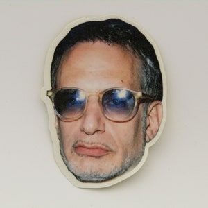 May include: A die-cut sticker featuring a man's face with sunglasses. The man has short, dark hair and a goatee. The sunglasses have a light-colored frame and blue lenses. The sticker is on a white background.