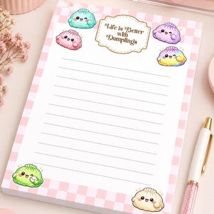 May include: A rectangular notepad featuring a light pink and white chequered border. The top centre displays a decorative label with the text "Life is Better with Dumplings." Several colourful, illustrated cartoon dumplings with faces are scattered around the text and at the bottom corners. The centre of the notepad contains horizontal lines for writing.