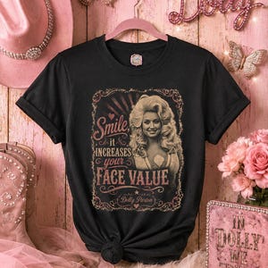 May include: A black short-sleeved t-shirt featuring a graphic print of Dolly Parton with the text "Smile it INCREASES your FACE VALUE" and "Dolly Parton" in a decorative frame. The shirt has a crew neckline and is displayed on a pink hanger.