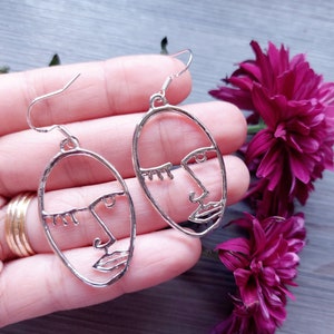 Può includere: Un paio di orecchini d'argento con un design a contorno di viso. Gli orecchini pendono dalla mano di una persona.