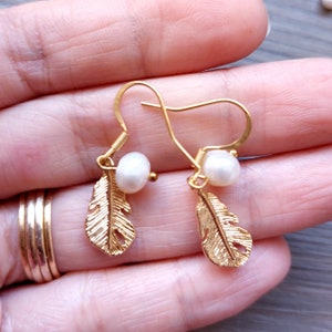May include: Gold feather earrings with a white pearl dangle. The earrings have a simple hook closure.