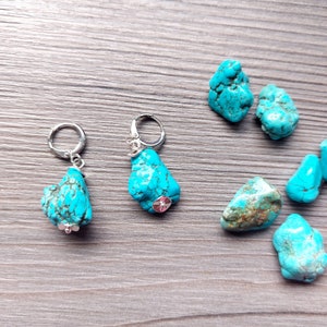 May include: A pair of turquoise stone earrings with silver hoops. The earrings are displayed on a wooden surface with several other turquoise stones.