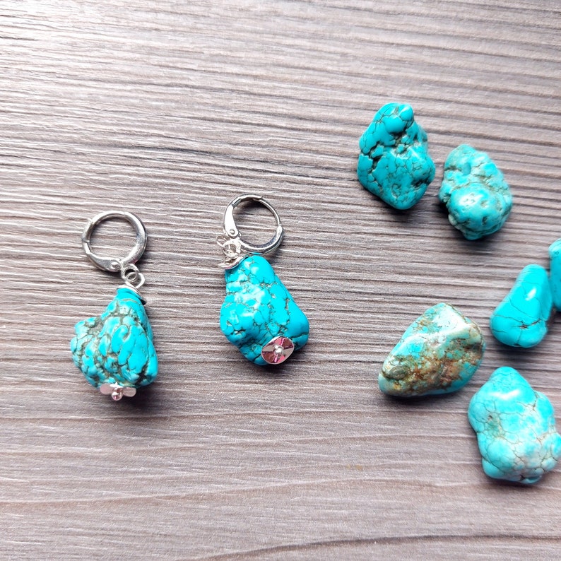 May include: A pair of turquoise stone earrings with silver hoops. The earrings are displayed on a wooden surface with several other turquoise stones.