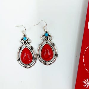 May include: A pair of silver dangle earrings with red and blue gemstone accents. The earrings feature a teardrop-shaped red stone surrounded by a silver frame.