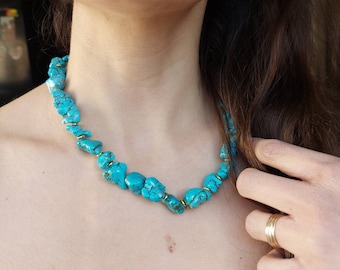 Puede incluir: Un collar de piedras turquesas con detalles dorados. El collar presenta piedras turquesas de forma irregular unidas con pequeños conectores dorados. El collar se muestra en el cuello de una persona.