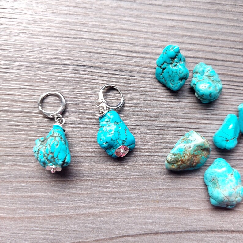 May include: Two pairs of turquoise earrings with silver hoops and turquoise stones. The earrings are displayed on a wooden surface with several turquoise stones.