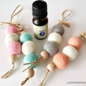 May include: Four felt ball car air fresheners with a wooden bead at the bottom and a small wooden peg at the top. Each air freshener has a different colour scheme: pink, white, and blue; grey, white, and peach; grey, white, and peach; and pink, white, and blue. A small bottle of lavender essential oil is in the centre of the image.