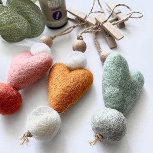 Op de afbeelding: Drie vilten hartvormige ornamenten met houten kralen en witte vilten ballen. De harten zijn roze, oranje en groen. De ornamenten hangen aan een touwtje met houten wasknijpers.