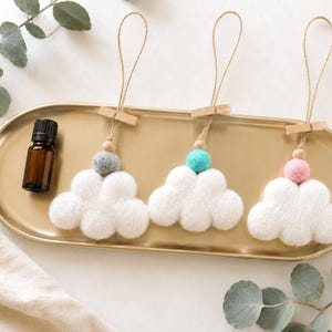 May include: Three white felt cloud ornaments with jute hanging loops, each featuring a small wooden bead and a single felt ball in gray, turquoise, or pink. A small amber glass bottle with a black cap is positioned next to the cloud ornaments.