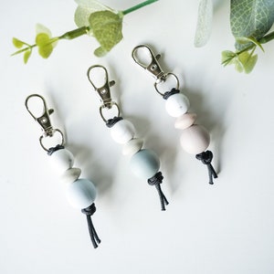 May include: Three keychains with silver clasps and black cords. Each keychain has three beads: a white bead, a light blue bead, and a light pink bead.
