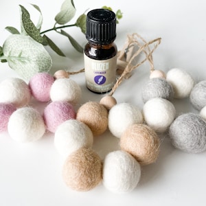 May include: Three felt ball garlands in pink, beige, and gray with a small bottle of lavender essential oil. The garlands are strung on twine and have wooden beads.