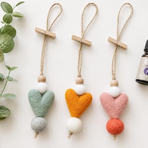 Può includere: Tre ornamenti pendenti con cuori in feltro color verde salvia, giallo senape e rosa antico. Ogni cuore è decorato con perline bianche e in legno naturale, sospeso tramite uno spago con una piccola molletta in legno. Un flacone di olio di lavanda Bluzen.