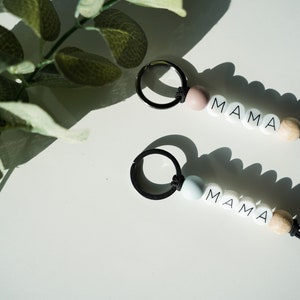 May include: Two keyrings with the word "MAMA" spelled out in white letter beads. One keyring has a pink bead, the other a light blue bead. Both have a wooden bead and a black keyring.