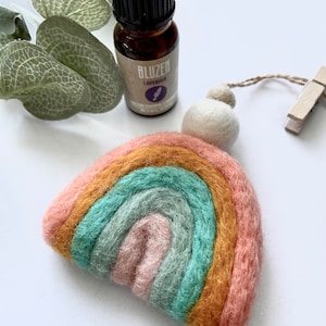 May include: A felt rainbow ornament with a white pom-pom and a wooden clothes peg. The ornament is hanging from a string. A small bottle of lavender essential oil is in the background.