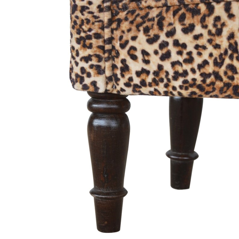 K&ouml;nnte beinhalten: Nahaufnahme der Beine eines Leopard-Muster-Aufbewahrungspuffs. Die Beine sind aus dunkelbraunem Holz und haben ein gedrehtes Design.