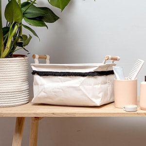 May include: A white canvas storage basket with black fringe trim and wooden handles. The basket is sitting on a wooden shelf with other bathroom items.