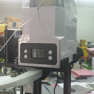 Può includere: Un porta-filamento bianco con un coperchio trasparente, attaccato a una stampante 3D. Il porta-filamento ha un display digitale che mostra "0004".