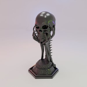 Puede incluir: Una escultura de calavera negra con manos esqueléticas sujetando su rostro. La calavera está posada sobre una base texturizada con una cola puntiaguda.