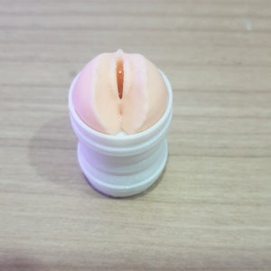 Pode incluir: Um recipiente de plástico branco com um inserto de silicone rosa. O inserto tem uma superfície texturizada com uma abertura elevada.