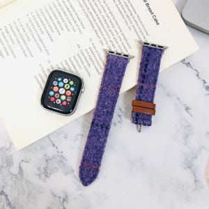 May include: A close-up of an Apple Watch with a black case and a purple tweed watch band with brown leather accents. The watch face displays colorful app icons. A recipe book is in the background.