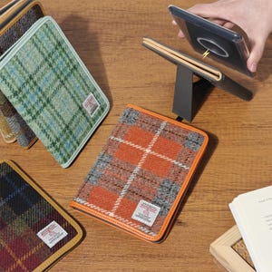 Peut inclure: Une collection de housses rectangulaires pour tablette ou carnet en laine Harris Tweed avec divers motifs à carreaux orange, verts et bleu foncé. Chaque housse possède une étiquette blanche et un support intégré pour incliner l'appareil.