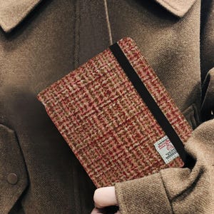 May include: A rectangular notebook with a red, brown, and white tweed cover. A black elastic band secures the notebook. A small label is visible on the bottom right corner. The notebook is held against a brown coat.